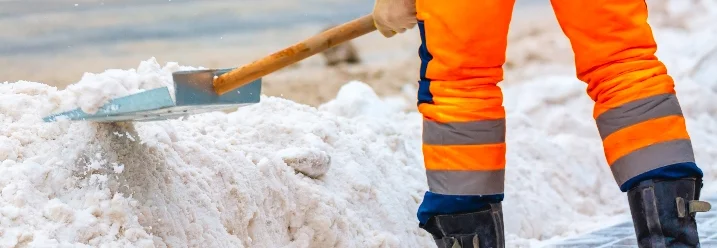 Person in oranger Arbeitskleidung räumt Schnee mit Schneeschieber