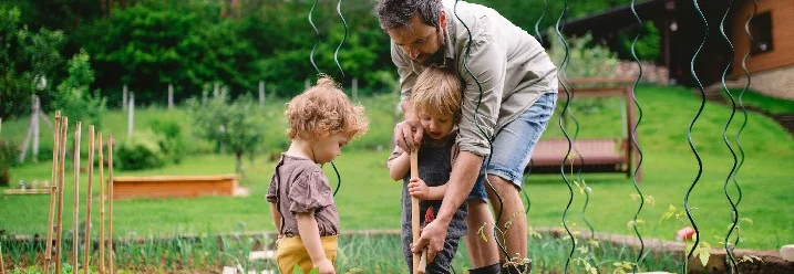 Erwachsener und zwei Kinder pflanzen mit einem Spaten etwas in einem Garten ein.