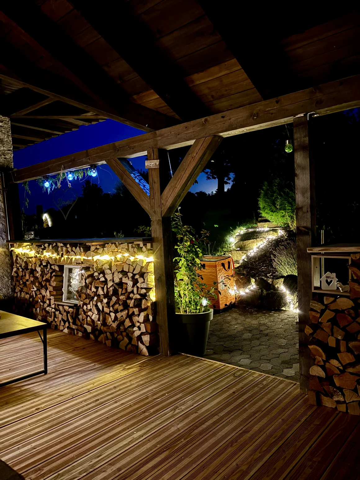 Eine beleuchtete Holzterrasse mit gestapeltem Brennholz bei Nacht.