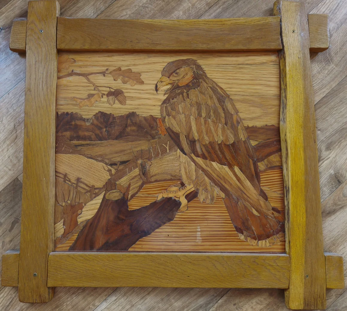 Gerahmte Holzintarsie eines Greifvogels auf einem Ast in einer Landschaft.