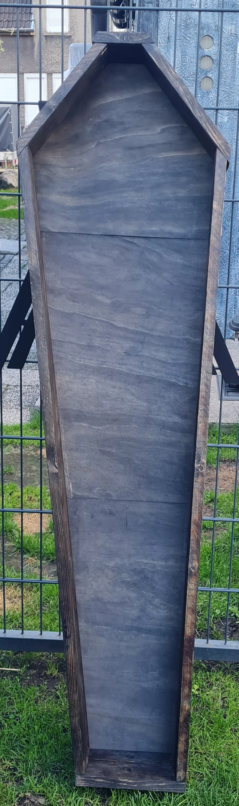 Sargförmiges Regal aus dunklem Holz, das an einem Zaun lehnt.