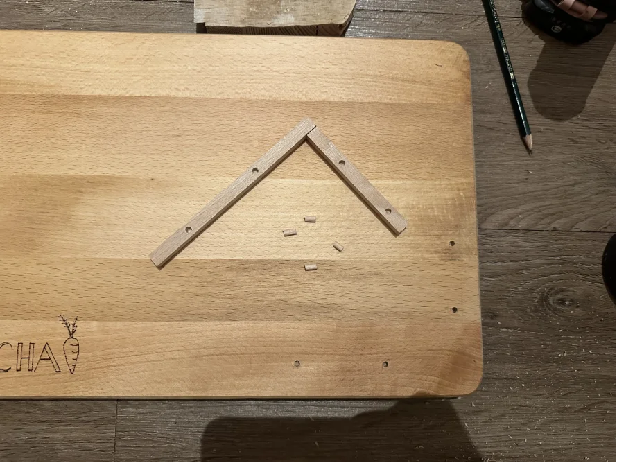 Zwei Holzlatten bilden ein Dreieck auf einem Holzbrett. Daneben liegen Holzdübel.