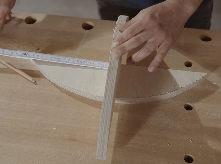 Zwei Hände richten ein gebogenes Stück Holz auf einer Werkbank aus. Ein Zollstock und ein Bleistift liegen daneben.