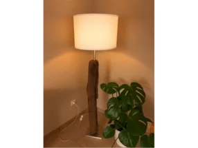 Eine beleuchtete Stehlampe mit einem Fuß aus Holz neben einer Zimmerpflanze.