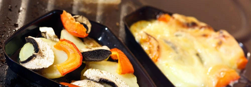 Zwei Raclette-Pfännchen mit Gemüse auf einem Grill, eines davon mit Käse überbacken.