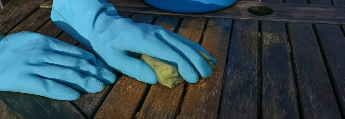 Eine Person reinigt einen Gartentisch aus Holz mit einem Schwamm.