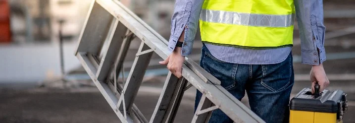 Person in Warnweste trägt Leiter und Werkzeugkasten.