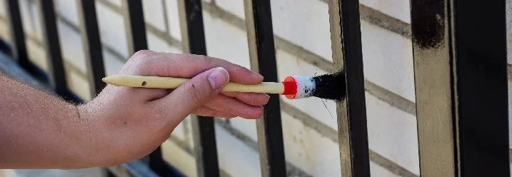 Person streicht Metallzaun mit Pinsel schwarz.