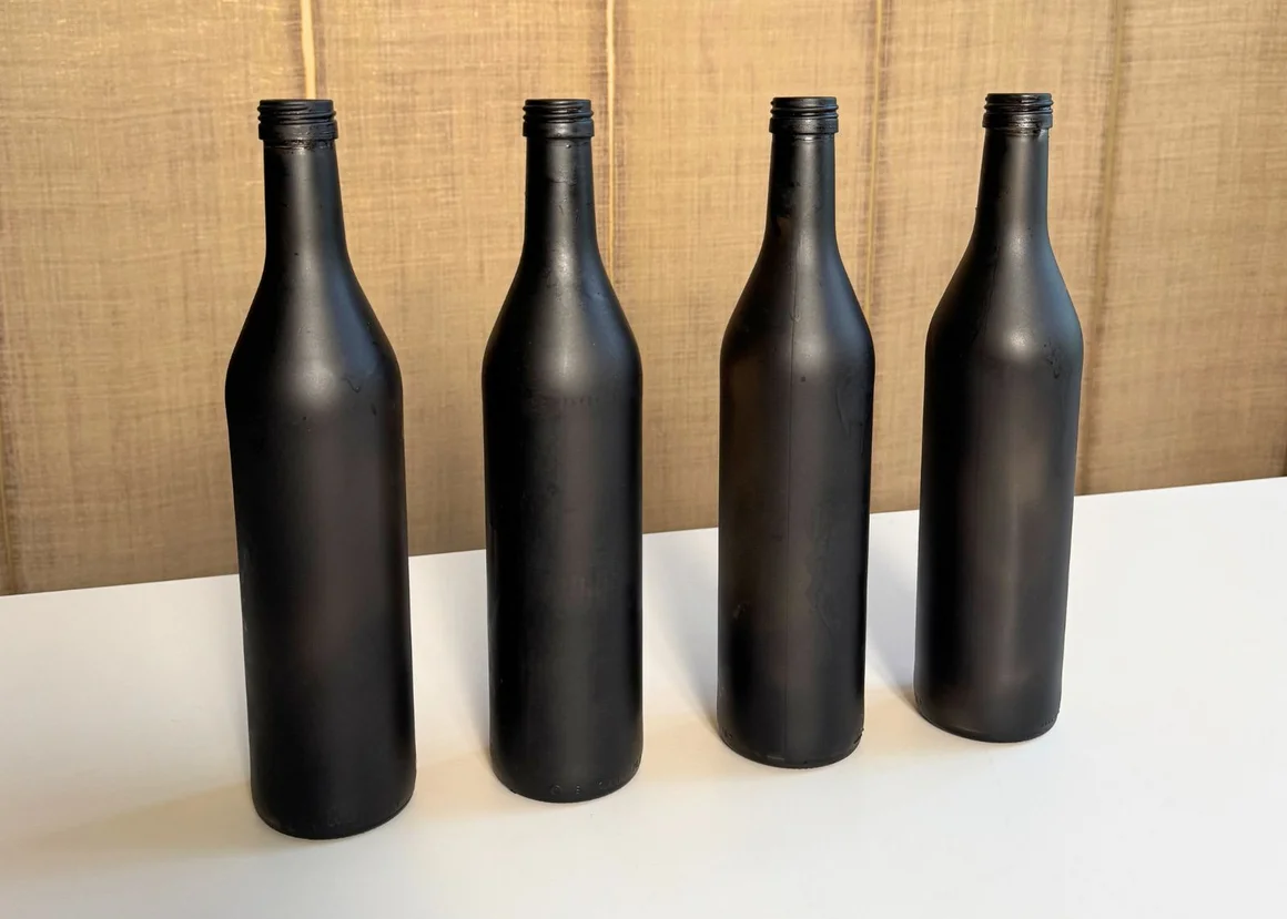 Vier mattschwarze Flaschen stehen in einer Reihe auf einer weißen Oberfläche.